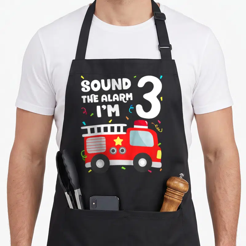 Toddler firefighter truck apron - Red fire truck with Sound The Alarm I'm 3 text and confetti — shown on a black cotton canvas apron against a white background