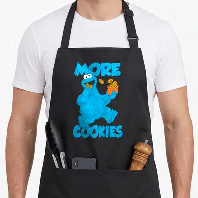 Longsleeve cookies apron - Blue character eating cookie holding orange bag with text MORE COOKIES — shown on a black cotton canvas apron against a white background