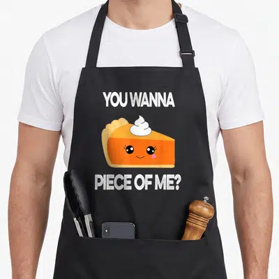Pumpkin thanksgiving apron - Cartoon orange pie slice with face and You Wanna Piece of Me text — shown on a black cotton canvas apron against a white background