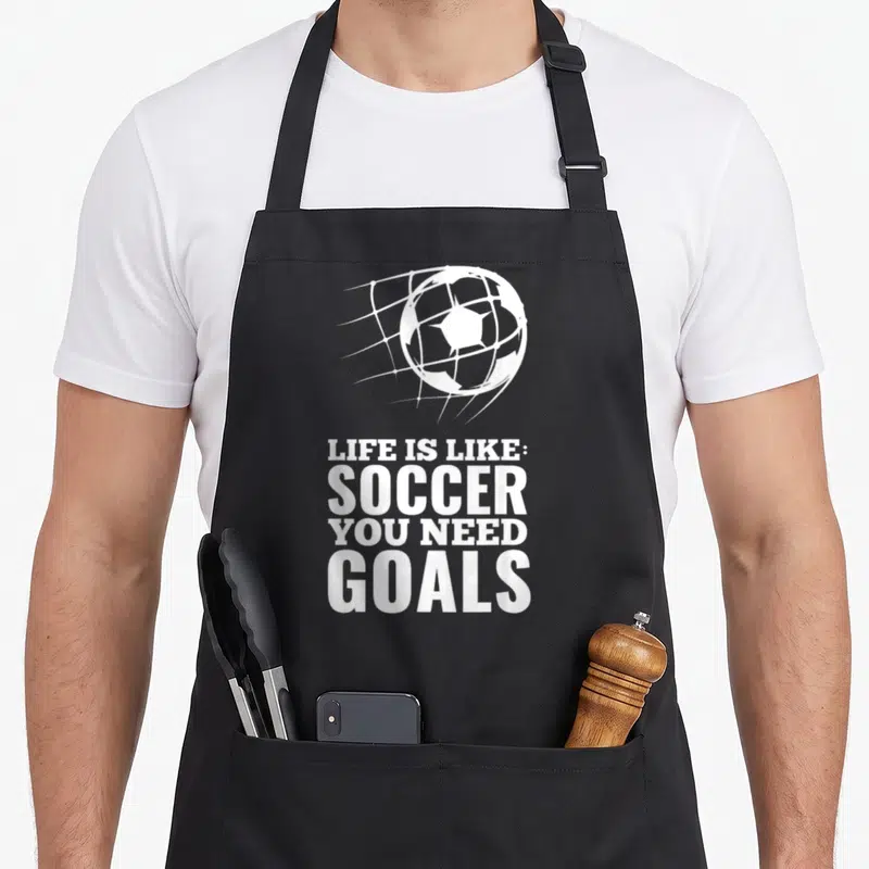 Goal funny soccer apron - White soccer ball hitting net above Life Is Like Soccer You Need Goals text — shown on a black cotton canvas apron against a white background