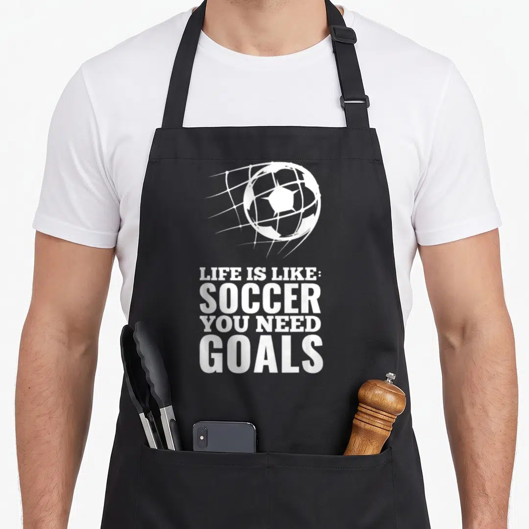 Goal funny soccer apron - White soccer ball hitting net above Life Is Like Soccer You Need Goals text — shown on a black cotton canvas apron against a white background