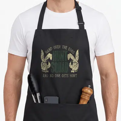 Rabbit chicken egg apron - Tan bunny silhouettes holding chickens flanking dark green Hunt Squad text in distressed — shown on a black cotton canvas apron against a white background