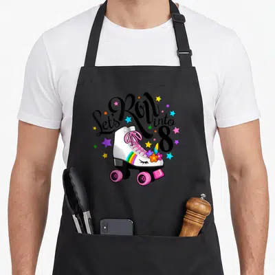 8th roller birthday apron - White roller skate with pink laces and Let's Roll into 8 text surrounded by colorful stars — shown on a black cotton canvas apron against a white background