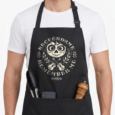 Coco cocoon apron - Beige sugar skull with two crossed guitars and RECUÉRDAME REMEMBER ME text in line art — shown on a black cotton canvas apron against a white background
