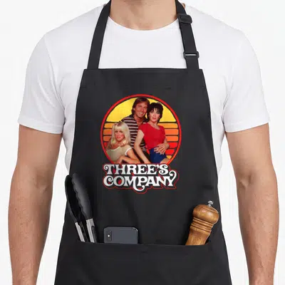 Three 1980s apron - Three people in front of yellow orange sun with white text reading THREE'S COMPANY below — shown on a black cotton canvas apron against a white background