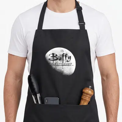 Opening slayer apron - White cratered moon with grey text reading Buffy the vampire slayer logo — shown on a black cotton canvas apron against a white background