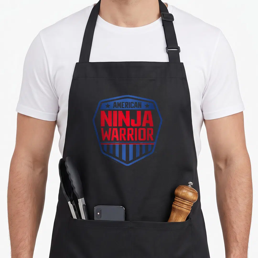 Ninja warrior apron - Blue hexagon logo with red AMERICAN NINJA WARRIOR text, two stars, and vertical stripes — shown on a black cotton canvas apron against a white background
