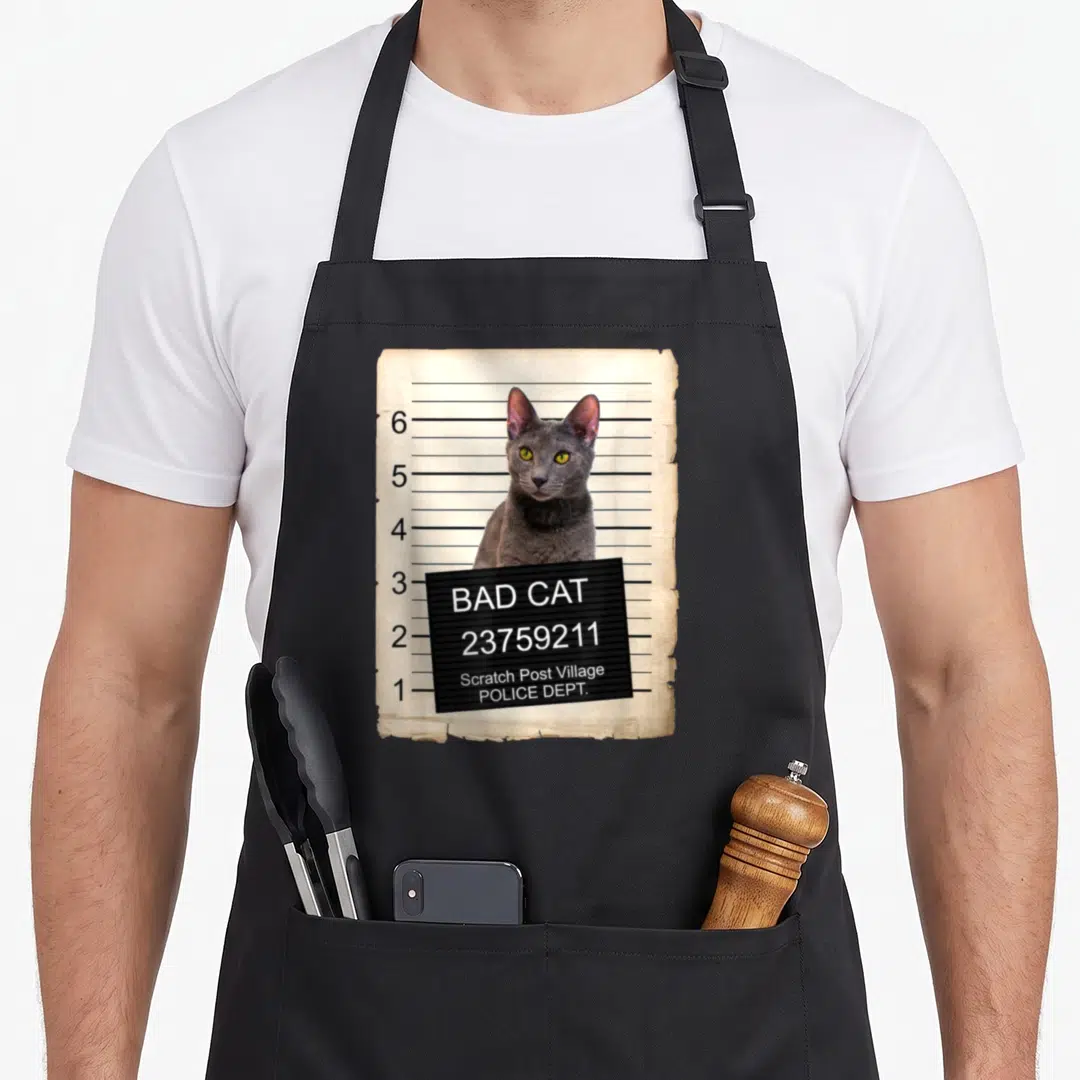 Kitten russian aprons - Grey cat mugshot on aged paper with black board reading BAD CAT Scratch Post Village POLICE DEPT — shown on a black cotton canvas apron against a white background