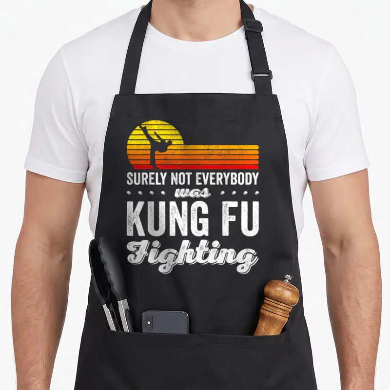 Surely kung apron - Kicking silhouette against retro striped sun with surely not everybody was kung fu fighting text — shown on a black cotton canvas apron against a white background