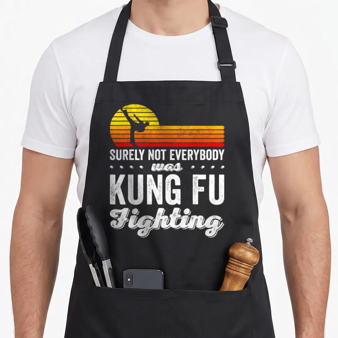 Surely kung apron - Kicking silhouette against retro striped sun with surely not everybody was kung fu fighting text — shown on a black cotton canvas apron against a white background