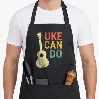Uke guitar ukulele apron - Distressed ukulele silhouette next to stacked UKE CAN DO IT text in multi-colors — shown on a black cotton canvas apron against a white background