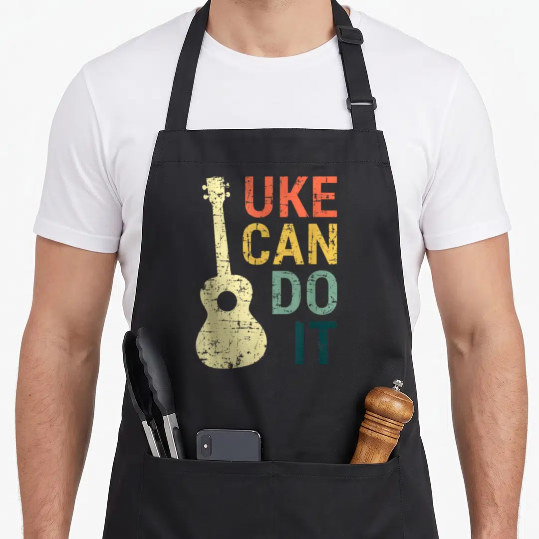 Uke guitar ukulele apron - Distressed ukulele silhouette next to stacked UKE CAN DO IT text in multi-colors — shown on a black cotton canvas apron against a white background