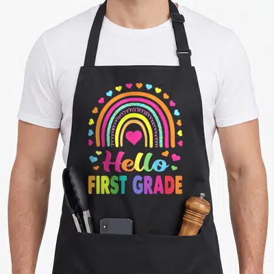 Hello 1st teacher apron - Rainbow with central heart and scattered hearts over Hello FIRST GRADE text in pink yellow — shown on a black cotton canvas apron against a white background
