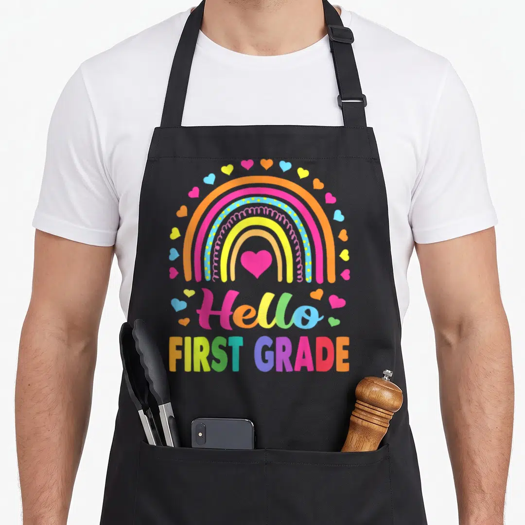 Hello 1st teacher apron - Rainbow with central heart and scattered hearts over Hello FIRST GRADE text in pink yellow — shown on a black cotton canvas apron against a white background