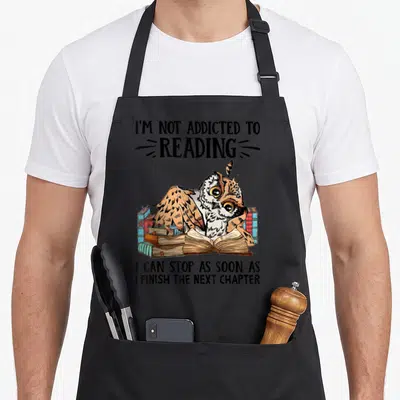 Addicted owl apron - Cartoon owl among blue red and brown books with text I'm not addicted to reading — shown on a black cotton canvas apron against a white background