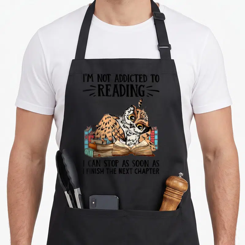 Addicted owl apron - Cartoon owl among blue red and brown books with text I'm not addicted to reading — shown on a black cotton canvas apron against a white background