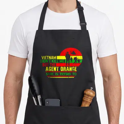 Agent retired veteran apron - Vietnam Agent Orange text with red yellow green striped circle and soldier silhouettes — shown on a black cotton canvas apron against a white background