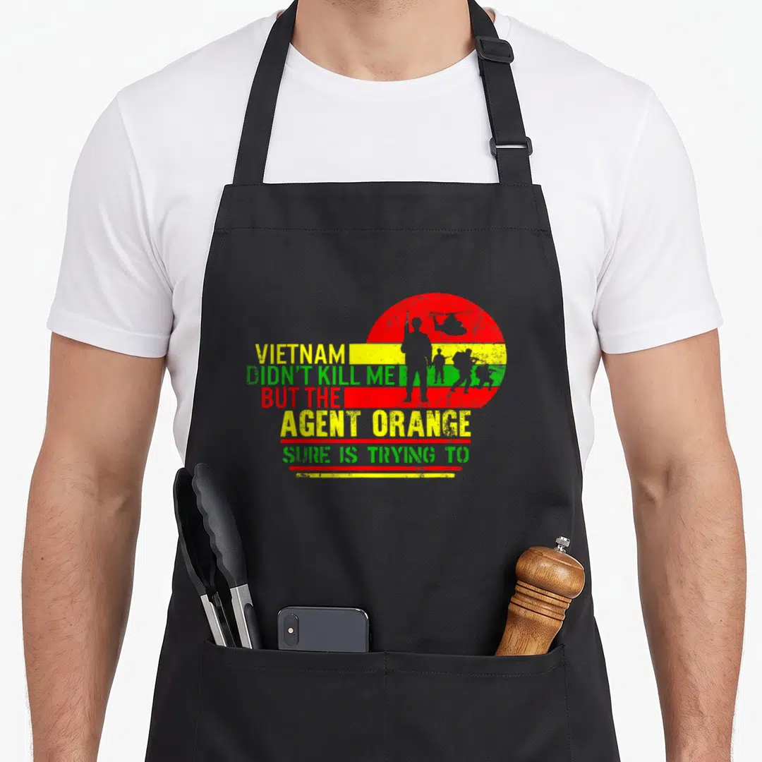 Agent retired veteran apron - Vietnam Agent Orange text with red yellow green striped circle and soldier silhouettes — shown on a black cotton canvas apron against a white background