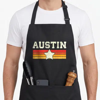 Funny austin texas aprons - Distressed AUSTIN text over red orange yellow stripes with a centered white star — shown on a black cotton canvas apron against a white background