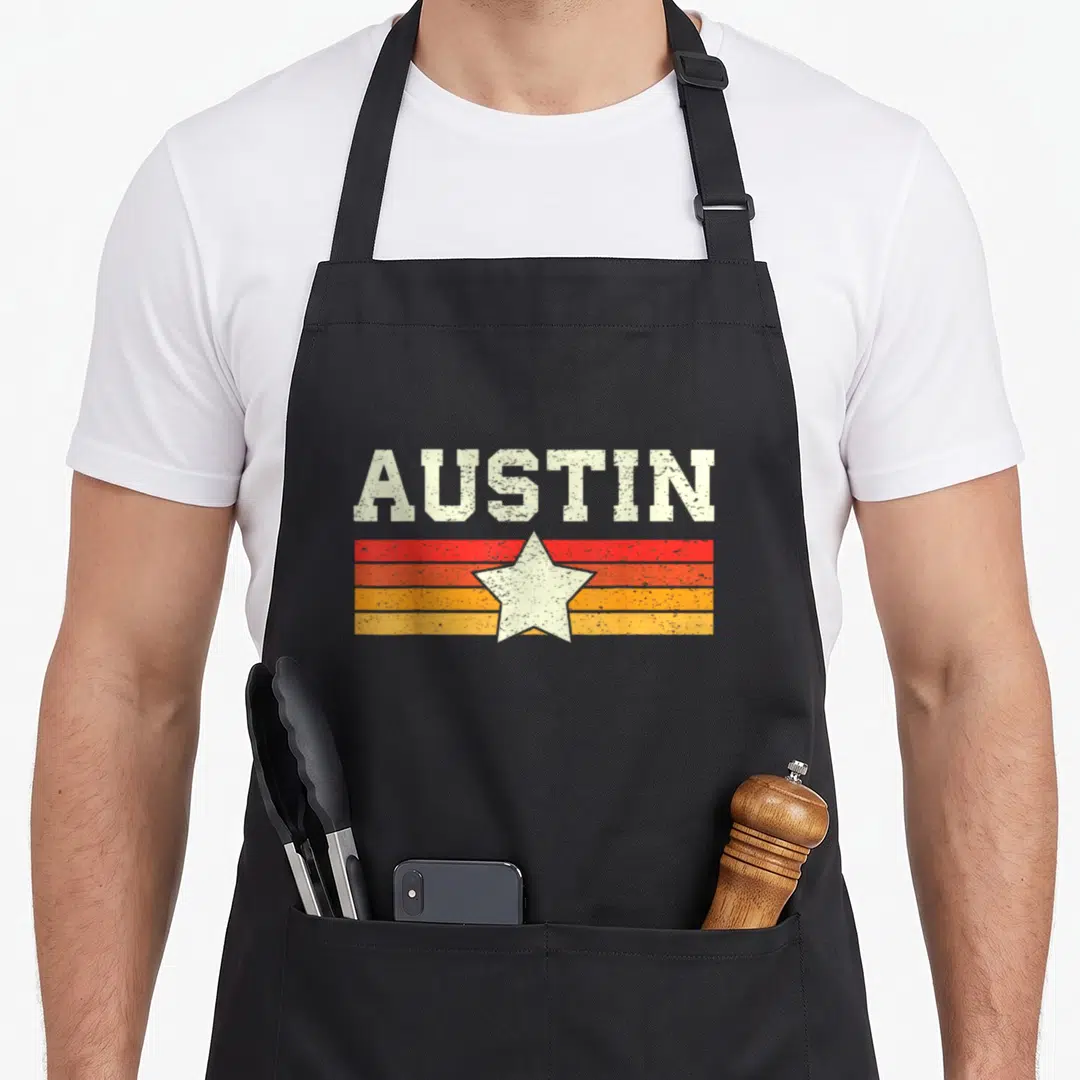 Funny austin texas aprons - Distressed AUSTIN text over red orange yellow stripes with a centered white star — shown on a black cotton canvas apron against a white background