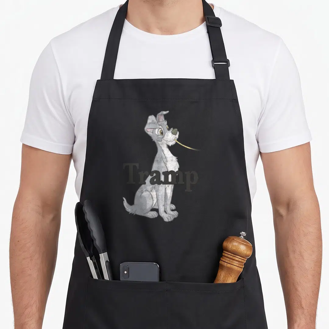 Funny spaghetti lady and the tramp apron - Grey cartoon dog holding yellow strand with Tramp text across body — shown on a black cotton canvas apron against a white background