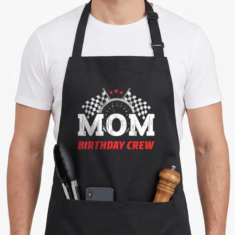 Theme crew aprons - MOM BIRTHDAY CREW text over speedometer with checkered flags and stars — shown on a black cotton canvas apron against a white background