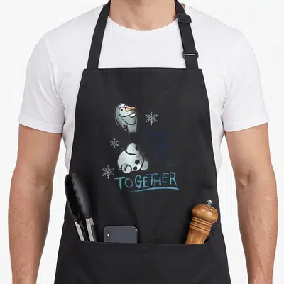 Smiling frozen apron - Cartoon snowman with text TRYING To KEEP IT TOGETHER and three snowflakes — shown on a black cotton canvas apron against a white background
