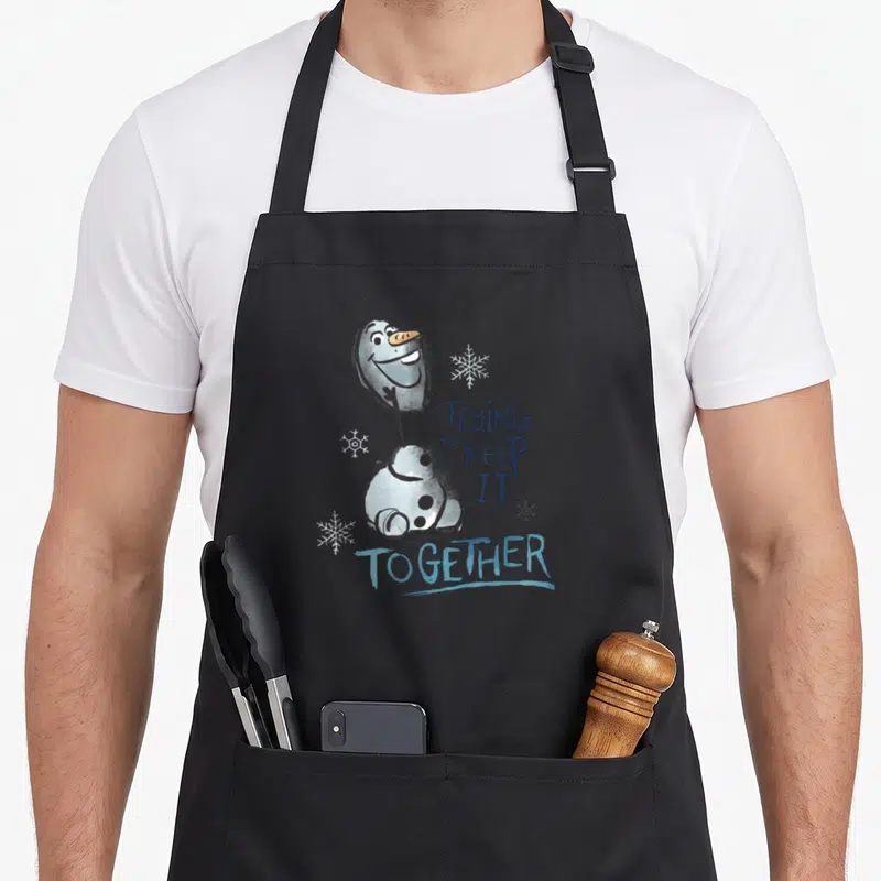 Smiling frozen apron - Cartoon snowman with text TRYING To KEEP IT TOGETHER and three snowflakes — shown on a black cotton canvas apron against a white background