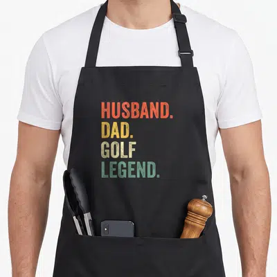 Husband golf apron - Stacked HUSBAND. DAD. GOLF LEGEND. text in orange, yellow, beige, and green distressed font — shown on a black cotton canvas apron against a white background