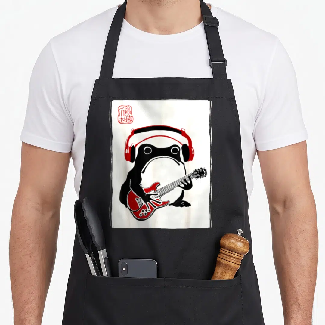 Grumpy kawaii aprons - Black frog wearing red headphones playing red electric guitar on white background with red seal — shown on a black cotton canvas apron against a white background