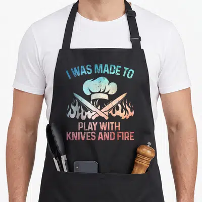 General culinary aprons - Gradient chef hat, crossed knives, flames, and text I WAS MADE TO PLAY WITH KNIVES AND FIRE — shown on a black cotton canvas apron against a white background