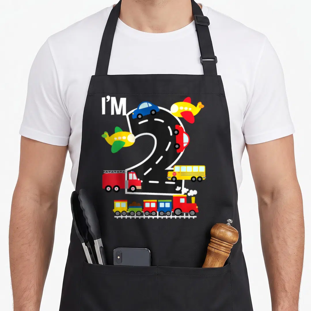 Bday truck apron - I'M 2 road graphic with cars, planes, fire truck, bus, and toy train in red blue yellow green — shown on a black cotton canvas apron against a white background