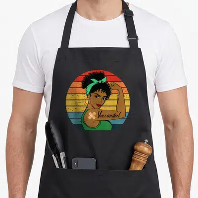 Afro pro apron - Woman flexing arm with Vaccinated text and bandages on striped background — shown on a black cotton canvas apron against a white background