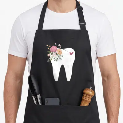 Oral dentist apron - White tooth graphic with pink and orange flower cluster and small heart on right side — shown on a black cotton canvas apron against a white background