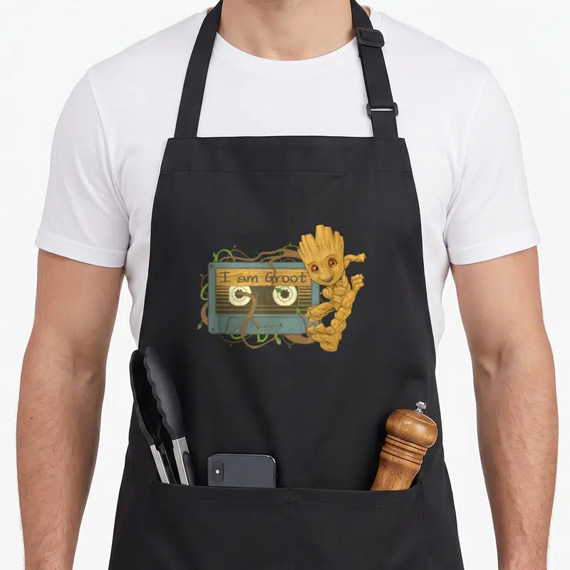 Guardian tapes lover apron - Baby Groot stands next to a teal cassette tape with vines and text reading I am Groot — shown on a black cotton canvas apron against a white background