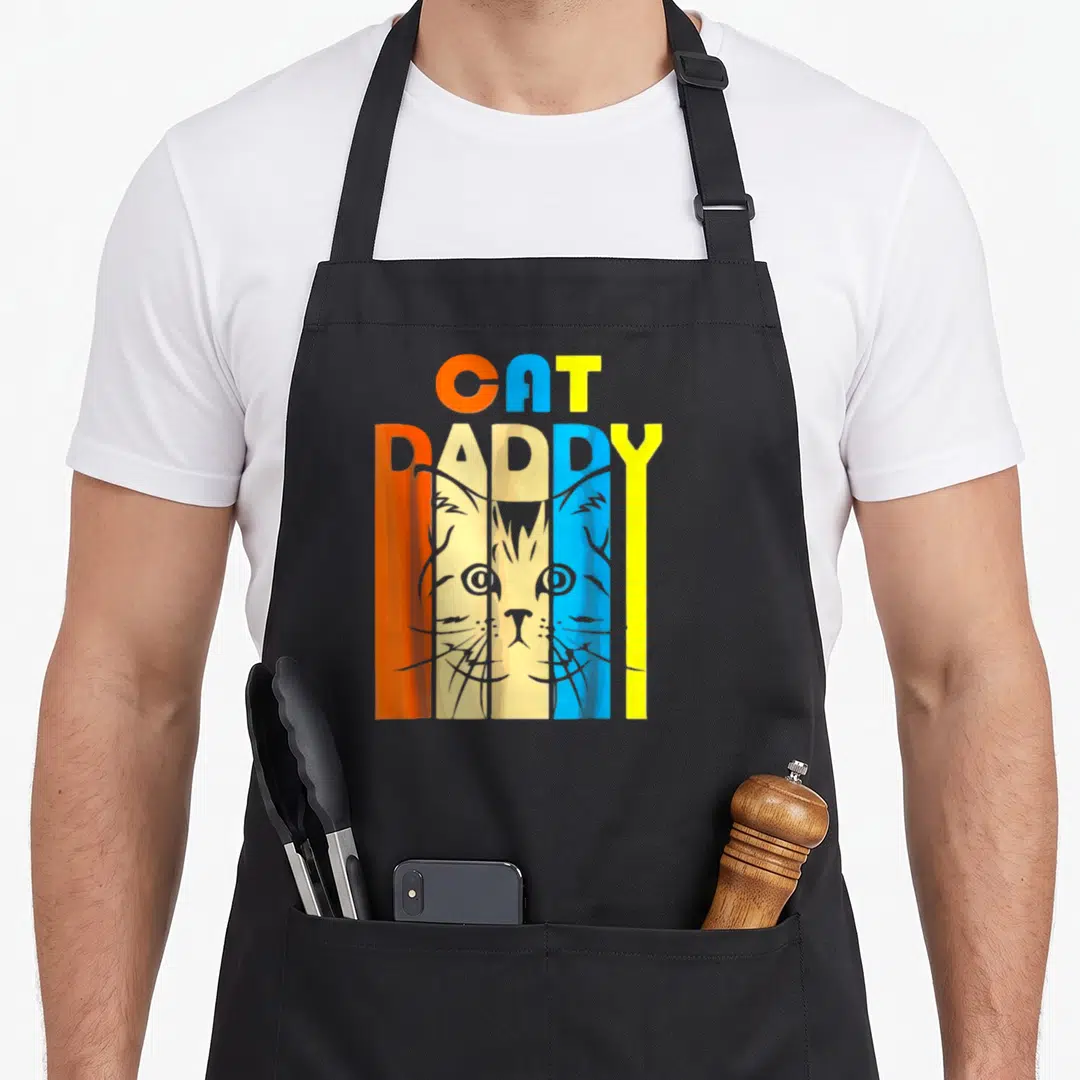 Cat daddy aprons - Retro CAT DADDY text with cat face behind vertical letters in orange beige blue yellow — shown on a black cotton canvas apron against a white background