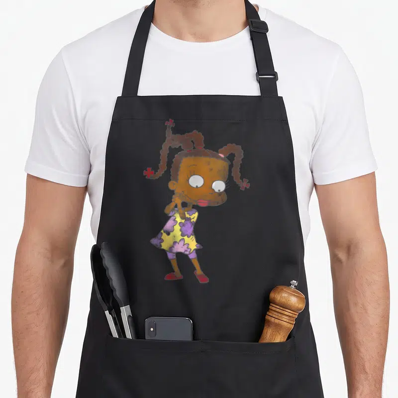 Susie watercolor apron - Cartoon girl with brown pigtails, yellow dress with purple flowers, and red shoes standing — shown on a black cotton canvas apron against a white background