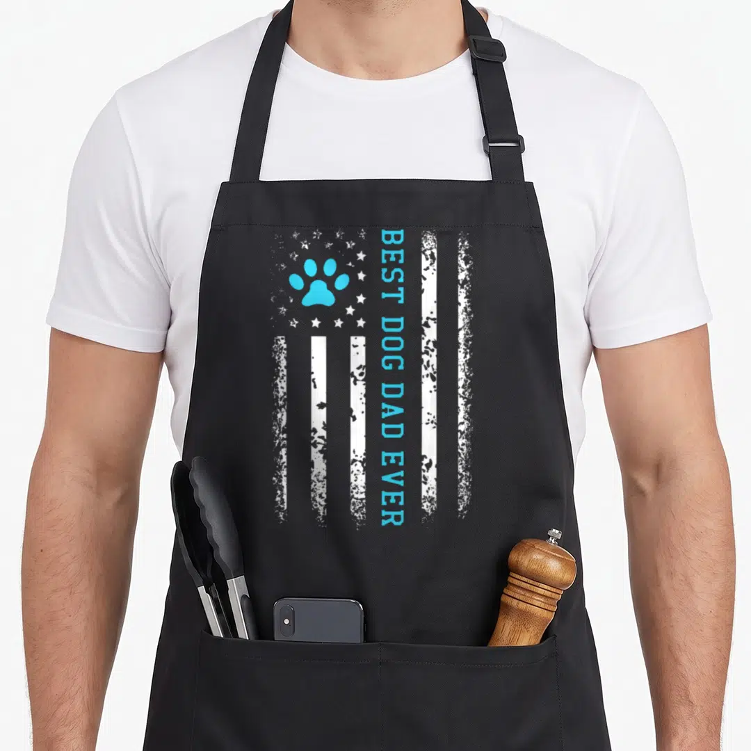 Dog american flag apron - Distressed white flag with light blue paw print and vertical BEST DOG DAD EVER text — shown on a black cotton canvas apron against a white background