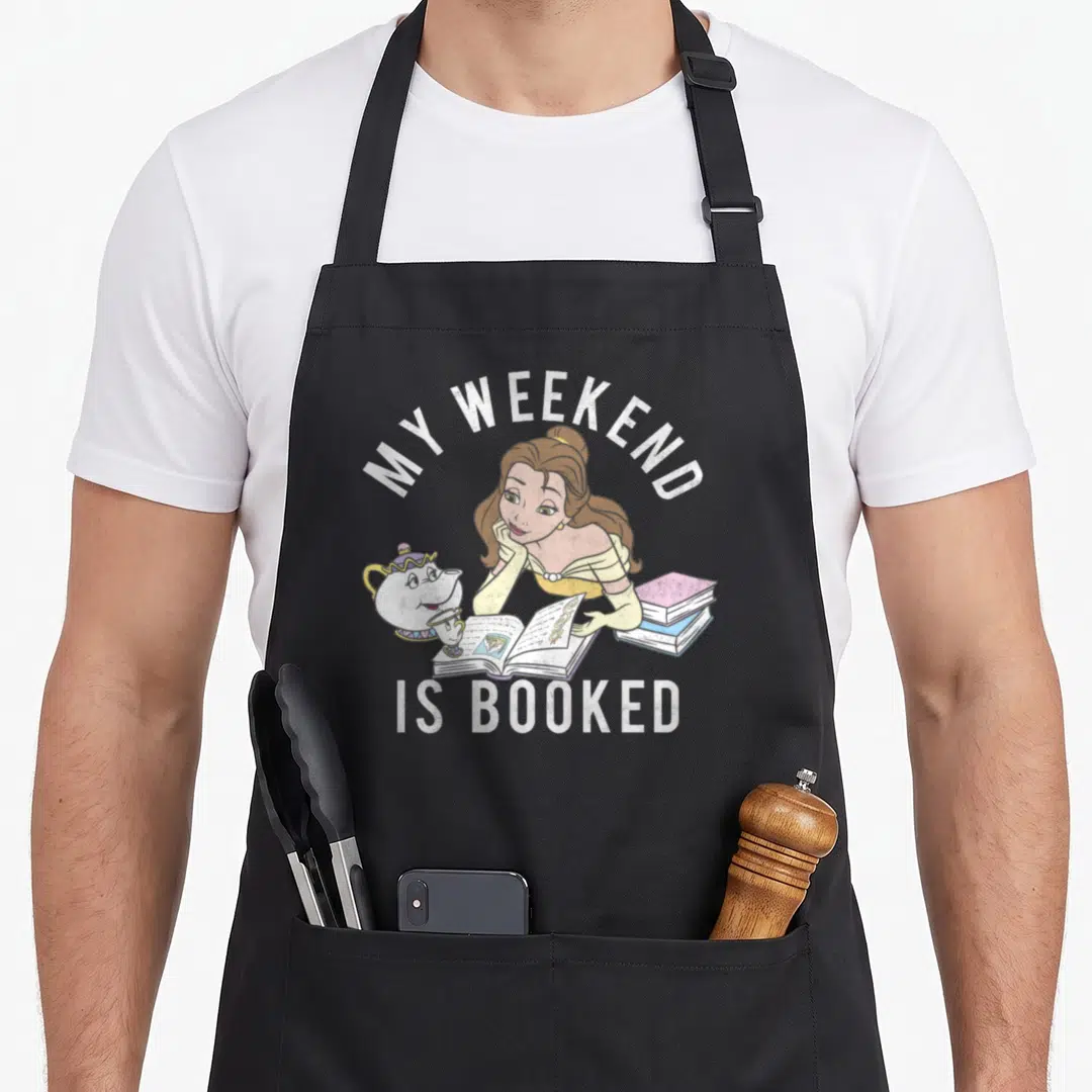 Weekend booked belle apron - Belle reading book with teapot and stacked books, MY WEEKEND IS BOOKED text in cartoon — shown on a black cotton canvas apron against a white background