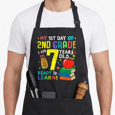 School 2nd apron - Rainbow text MY 1ST DAY OF 2ND GRADE around yellow number 7 with pencil and books — shown on a black cotton canvas apron against a white background
