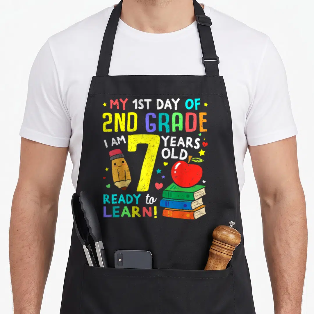 School 2nd apron - Rainbow text MY 1ST DAY OF 2ND GRADE around yellow number 7 with pencil and books — shown on a black cotton canvas apron against a white background