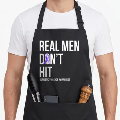 Hit domestic aprons - White stacked text reading Real Men Don't Hit with purple ribbon inside the O — shown on a black cotton canvas apron against a white background