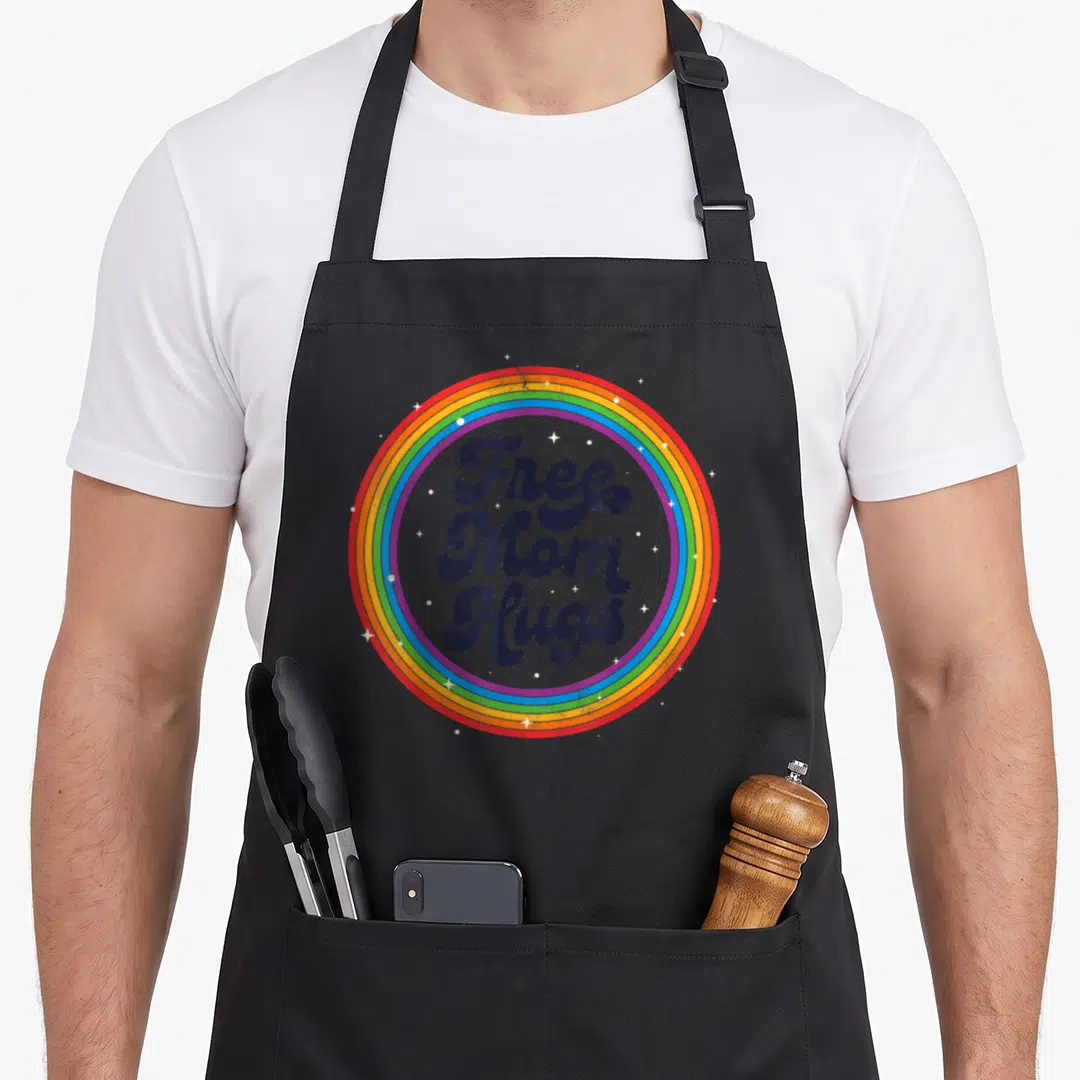 Mother gay pride apron - Free Mom Hugs text in retro font inside rainbow circle with white stars — shown on a black cotton canvas apron against a white background