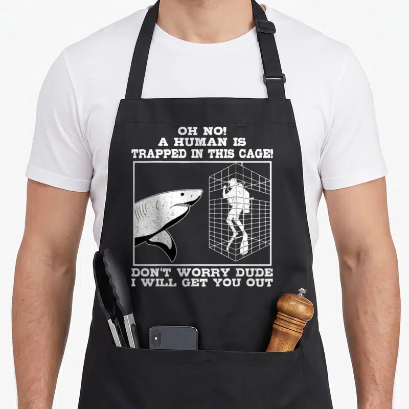 Shark predator apron - Distressed text above and below a shark facing a diver in a cage — shown on a black cotton canvas apron against a white background