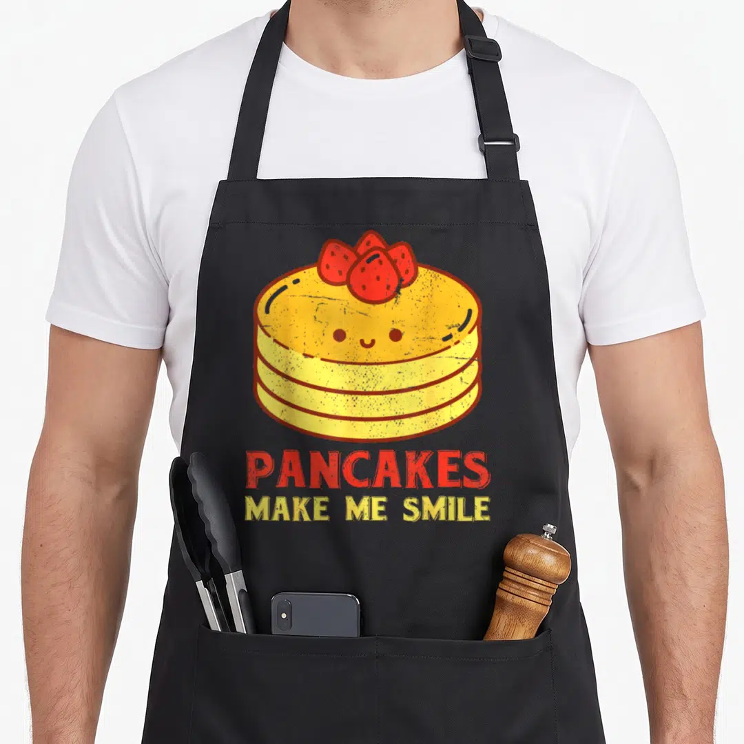 Food pancake apron - Cartoon yellow pancake stack with red strawberries and red and yellow text reading PANCAKES MAKE — shown on a black cotton canvas apron against a white background