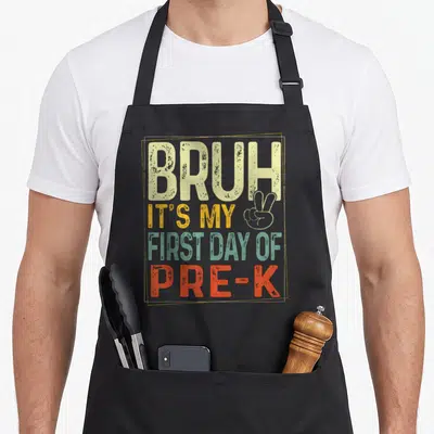 Bruh pre k apron - Stacked distressed text reading Bruh Its My First Day Of Pre-K in beige orange green and red — shown on a black cotton canvas apron against a white background