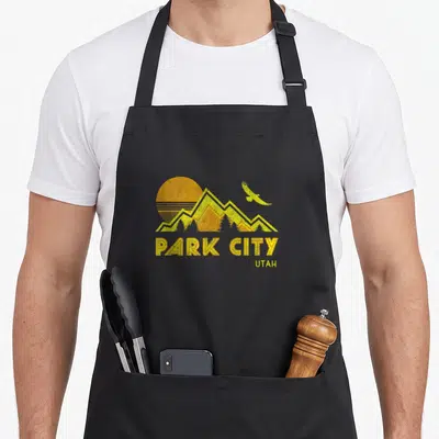 Park utah distressed apron - Yellow sun, mountains, and flying eagle above PARK CITY UTAH text on black background — shown on a black cotton canvas apron against a white background