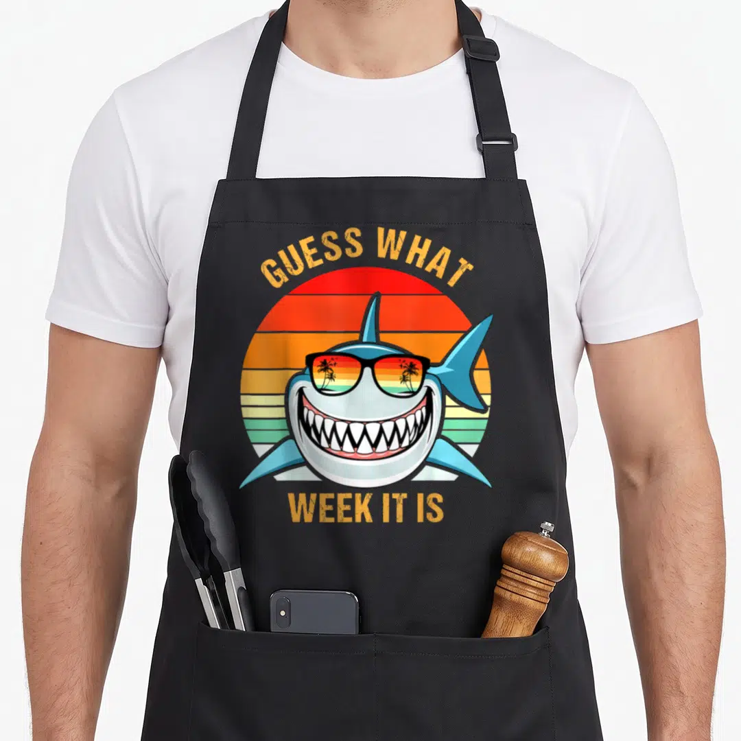 Guess i love marine apron - Cartoon shark head with sunglasses over striped retro sun with orange text — shown on a black cotton canvas apron against a white background