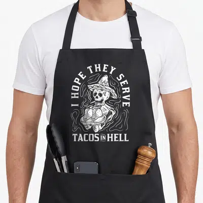Taco to serve man apron - White line-art skeleton in sombrero holding two tacos surrounded by smoke with text I HOPE — shown on a black cotton canvas apron against a white background