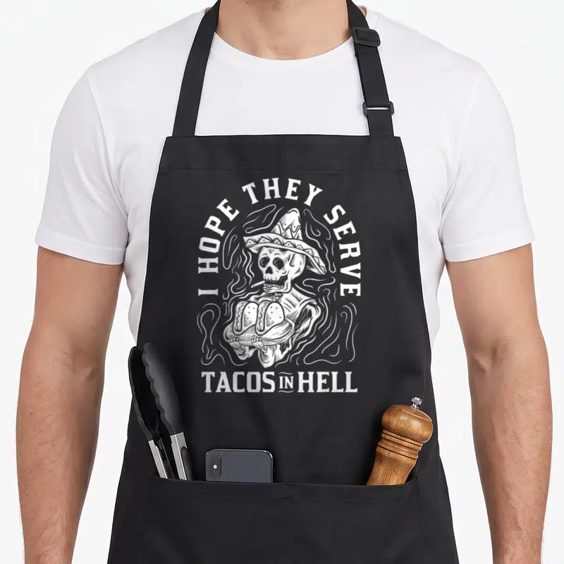 Taco to serve man apron - White line-art skeleton in sombrero holding two tacos surrounded by smoke with text I HOPE — shown on a black cotton canvas apron against a white background